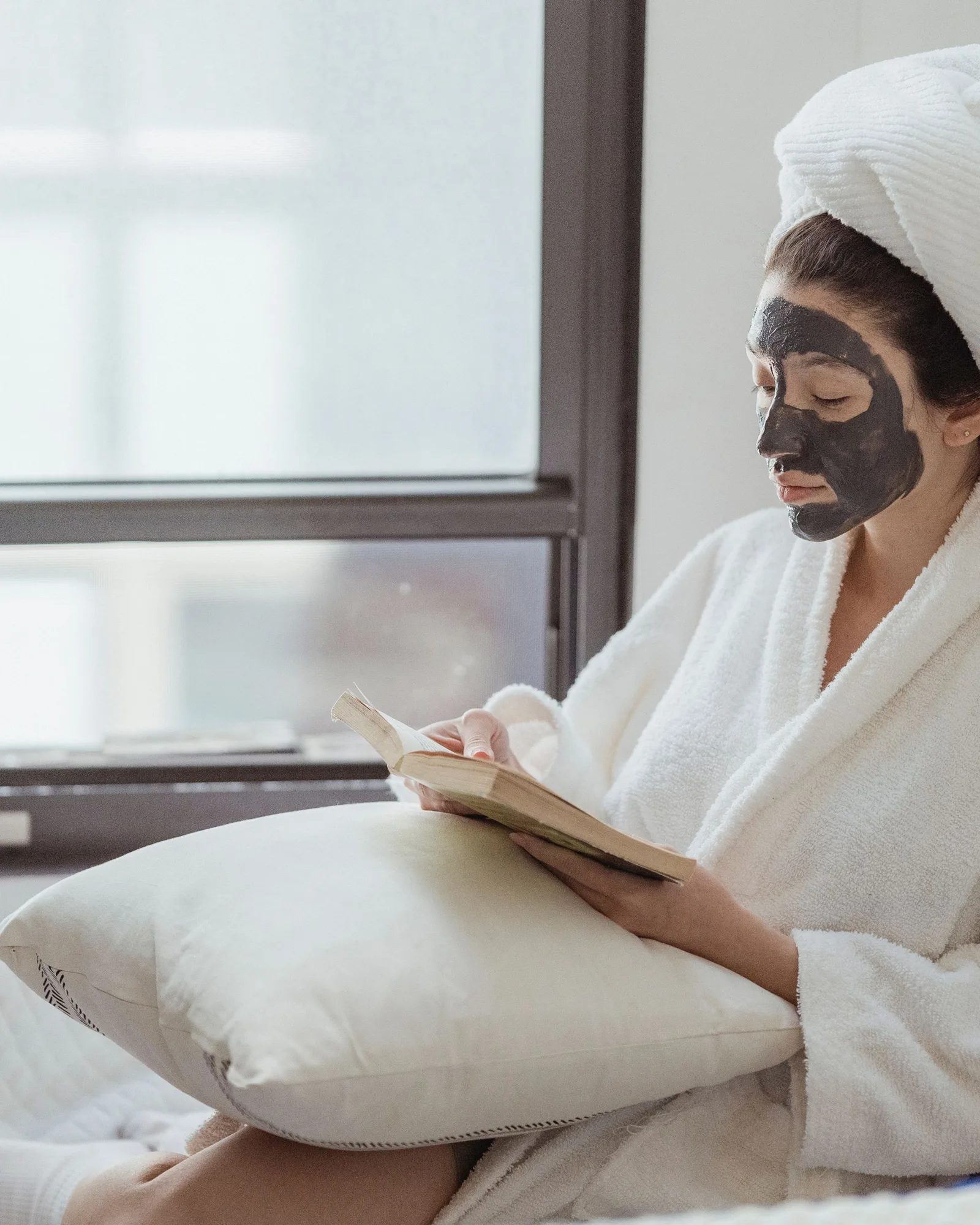 Une femme en peignoir lit le dos d’un tube de soin, des masques en sachet à la main